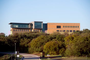 A modern, multi-story brick building with large windows stands behind a line of trees under a clear sky.