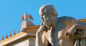 Stone statue of a seated philosopher with a contemplative pose, set against a blue sky. A building with a decorative eagle is visible in the background.