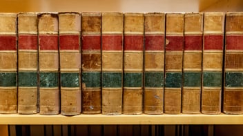 A row of vintage books with worn brown covers, featuring red and green bands, lined up on a wooden shelf.