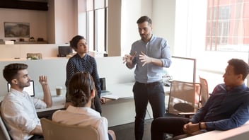 Five people in business attire are having a discussion in a modern office, with one person standing and speaking while the others sit and listen.