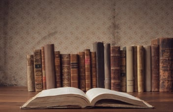 An open book lies on a table in front of a row of vintage books lined up against a patterned wallpaper.