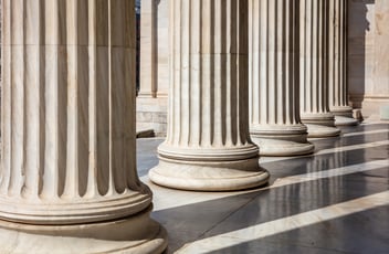 Row of large stone columns with intricate bases in a classical architectural style, casting shadows on a polished floor.