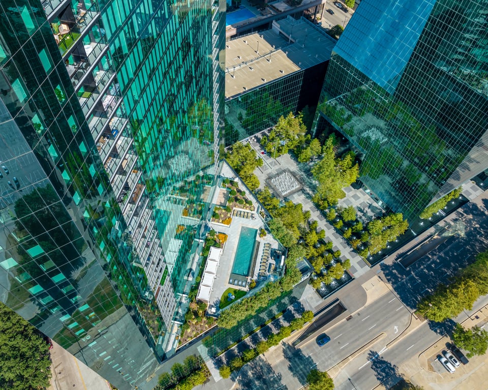 Aerial view of a modern glass skyscraper complex with a rooftop garden and pool area, surrounded by trees and streets.