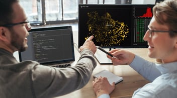 Two people at a desk discussing data analytics displayed on a monitor. One points at the screen showing yellow data points; a laptop with code is also visible.