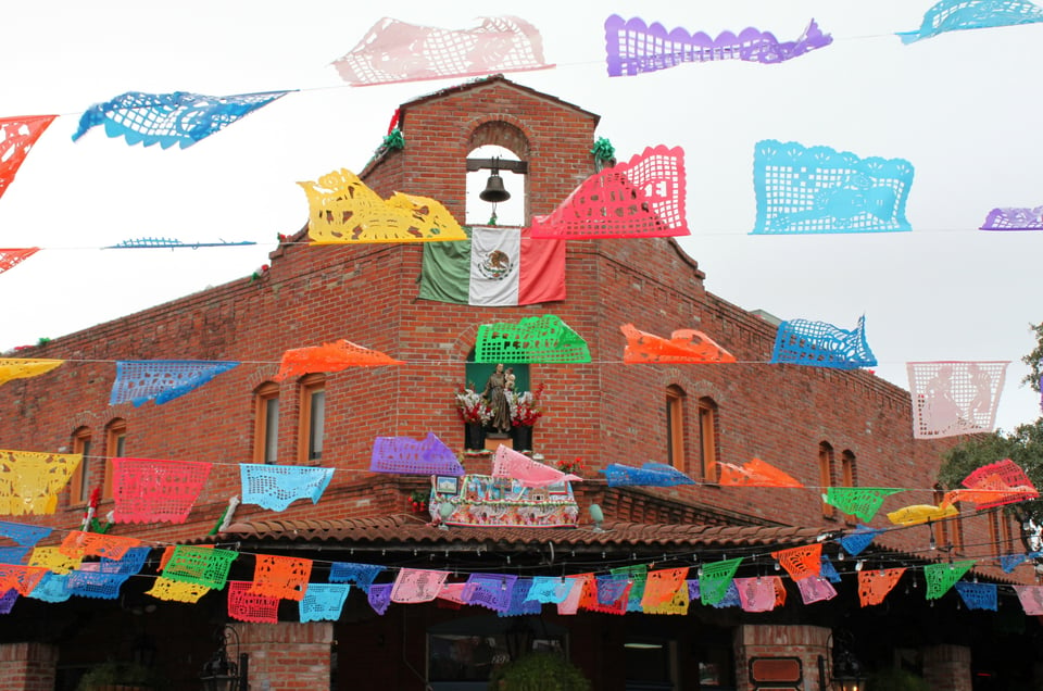 Colorful papel picado banners hang outside a brick building with a Mexican flag and bell on the roof.