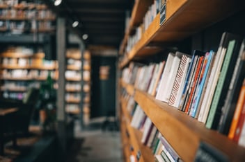 A library aisle with wooden shelves filled with books, extending into the distance. The background is slightly out of focus.