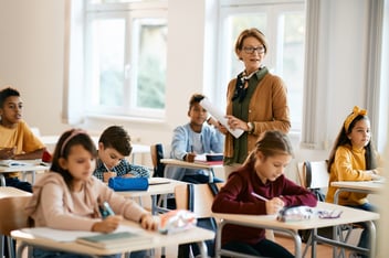 A teacher stands at the front of a classroom holding papers while students sit at desks writing and listening during a lesson.